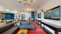 This single-story clubhouse in Oxford, Mississippi, offers a lively and eclectic communal environment designed for socializing and relaxation. The main lounge area is anchored by generously sized gray sectional sofas and a tufted ottoman, all set atop bold red carpeting that adds energy to the space. Geometric pendant chandeliers hang overhead, complementing the recessed lighting to create a warm, well-lit atmosphere. Decorative touches — including mounted antlers, framed artwork, and plaid accent chairs — give the space a distinctive, personality-driven character.

Large flat-screen televisions are mounted along the white brick accent wall, supported by dark console tables, making this area well-suited for watching sports or gatherings. Built-in bookshelves and reading nooks provide quieter corners for individual use. A decorative lattice room divider helps define separate zones within the open layout without interrupting the flow.

The kitchenette features bold green-painted walls, a striking floral wallpaper accent, white cabinetry with black countertops, open wood shelving, and light hardwood flooring — offering a bright and functional space for residents. Additional seating and workspace areas are visible throughout, rounding out this well-appointed and vibrantly designed clubhouse.