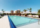 Located at 405 NE 2nd St in Fort Lauderdale, Florida, this amenity space delivers a comprehensive fitness and recreation experience across a single level. The fitness center is well-appointed with a wide range of commercial-grade equipment, including multiple treadmills, stair climbers, stationary bikes, and an extensive selection of strength-training machines designed to support full-body workouts. The interior features durable rubberized flooring, recessed lighting, and clean white walls that create a bright, functional environment. Large windows and glass double doors allow natural light to filter in throughout the space while providing views of the surrounding greenery and pool area, enhancing the overall workout atmosphere. Motivational artwork adorns the walls, adding character to the fitness environment. The outdoor area showcases a generously sized resort-style swimming pool with shimmering blue water framed by elegant tile detailing. Surrounded by lounge chairs, lush palm trees, and pergola structures, the pool deck provides a relaxing retreat with sweeping views of the Fort Lauderdale skyline. The combination of a fully equipped fitness center and an upscale outdoor pool area makes this space a standout amenity offering for its users.
