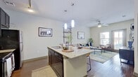 This single-floor residence is designed with a modern, open-concept layout that seamlessly connects the kitchen, dining, and living areas. The kitchen is a standout feature, equipped with dark espresso cabinetry reaching toward the ceiling, stainless steel appliances, a subway tile backsplash, and a central island topped with light granite countertops. Pendant lighting illuminates the island, which also functions as a casual dining and workspace with bar seating. Rich hardwood-style flooring flows throughout the entire unit, adding warmth and visual continuity. The living area is generously proportioned, bathed in natural light from a large window and a glass-paned exterior door that provides direct outdoor access. A ceiling fan adds comfort to this inviting space. The bedroom is a tranquil retreat, featuring dual windows that bring in abundant natural light, a ceiling fan, and a reach-in closet with shelving. The bathroom is well-appointed with dark cabinetry, granite countertops, a large mirror, and a full soaking tub and shower combination accented by large-format dark tile surround. Ample closet and storage space is present throughout, including a double-door closet near the entryway.
