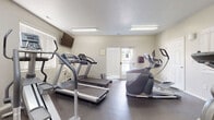 This fitness center offers a comprehensive workout environment designed to accommodate a variety of exercise routines. The space is laid out across a single floor and features durable rubber flooring throughout, providing both safety and comfort during workouts. The cardio section is well-stocked with multiple treadmills and elliptical trainers, allowing several users to exercise simultaneously. A stationary bike and a multi-function cable weight machine round out the strength and cross-training options, catering to a full range of fitness needs. Large wall-mounted mirrors line portions of the room, visually expanding the space and allowing users to monitor their form during exercise. A flat-screen TV is mounted on the wall, offering entertainment during workouts. Natural light filters in through multiple windows fitted with blinds, complementing the overhead fluorescent lighting to keep the space bright and energizing. A glass-paneled door provides direct access to the outdoors, enhancing ventilation and connectivity to the exterior. The neutral-toned walls and clean layout give the fitness center a functional, no-frills atmosphere focused entirely on supporting an active lifestyle.