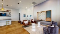 This single-floor amenity lounge is a thoughtfully designed social space that seamlessly blends modern sophistication with inviting warmth. The open layout is anchored by warm hardwood flooring that flows throughout, complemented by soaring ceilings that enhance the sense of spaciousness and airiness.

The lounge area features a curated mix of leather armchairs, plush velvet seating, and sculptural hourglass-shaped side tables arranged atop a neutral area rug, creating an inviting gathering spot. A large, framed oversized Scrabble board mounted on a dedicated accent wall serves as both a functional game feature and a striking focal point. Interactive kiosk stations are also integrated along one wall, adding a tech-forward element to the space.

The kitchen and bar area is equally impressive, centered around a generous island topped with a striking live-edge wood countertop. Woven rattan pendant lights hang overhead, casting warm ambient light. Open industrial-style shelving lines the white brick backsplash wall, displaying decorative accents alongside built-in appliances and light blue cabinetry below. Gold-accented bar stools provide casual seating at the island.

Floor-to-ceiling steel-framed windows allow abundant natural light to pour in, offering views of the surrounding exterior and connecting the interior to the outdoors. A billiards table rounds out the entertainment offerings, making this a versatile and well-appointed communal retreat.