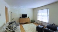 This single-floor space presents a thoughtfully arranged living area that balances simplicity with functionality. The room features classic parquet hardwood flooring, which adds warmth and a timeless character to the interior. A large window fitted with horizontal blinds allows generous natural light to filter into the space, brightening the neutral white walls and creating an airy, open atmosphere.

The living area is furnished with a comfortable sofa accented by patterned throw pillows, complemented by a plush white shag area rug that adds texture and coziness underfoot. A mid-century modern style wooden dresser serves as a media console, supporting a flat-screen television, and its warm wood tones echo the flooring below. A small green ottoman provides additional flexible seating or surface space nearby.

A baseboard heating unit runs along the exterior wall beneath the window, offering a practical and unobtrusive climate solution. Storage is subtly incorporated through a woven basket tucked into the corner, keeping the space organized without compromising its clean aesthetic. Overall, the space conveys a calm, livable ambiance with a modest yet welcoming character.