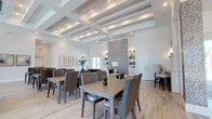 This single-story clubhouse is designed to impress, combining refined architectural details with a bright, welcoming atmosphere. The main gathering area showcases striking coffered ceilings with recessed lighting that casts a warm, even glow throughout the space. Light wood-style flooring runs underfoot, complementing the crisp white walls and lending the interior a clean, contemporary aesthetic.

The dining area is generously appointed with multiple tables and woven upholstered chairs, offering ample seating for social events and community gatherings. Adjacent to the dining space, a comfortable lounge area provides a more relaxed setting, anchored by a dramatic floor-to-ceiling textured accent wall that serves as a bold focal point. Built-in shelving units frame the lounge, offering both display and storage opportunities.

Natural light filters in through glass doors, enhancing the open and airy feel of the space. Thoughtful touches such as potted greenery and curated décor add warmth and life to the interior. The overall ambiance is polished yet approachable — a space that balances elegance with everyday functionality, making it well-suited for a wide range of community activities and social occasions.