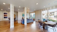 This single-story community clubhouse presents an open, well-appointed interior designed for both relaxation and social gathering. Throughout the space, warm wood-look flooring and signature tan-and-white striped wallcoverings create a cohesive, inviting aesthetic. Recessed lighting, complemented by a stylish multi-bulb chandelier, ensures the space is well-lit at all times.

The main lounge area anchors the layout, featuring a comfortable seating arrangement centered around a sofa and area rug, with a wall-mounted television and media console nearby. Large floor-to-ceiling windows and sliding glass doors flood this zone with natural light while offering views of the surrounding landscaped community grounds.

A casual dining nook with a round table and woven rattan chairs sits adjacent to the windows, providing an informal spot for residents to gather. The kitchenette area is equipped with stainless steel appliances, including a full-size refrigerator and dishwasher, wood-tone cabinetry, a sink, and open display shelving — ideal for hosting community events. A central island with bar-height seating offers additional dining space.

Completing the layout, a dedicated game area features a foosball table, adding a recreational element. White structural columns define zones within the open floor plan without interrupting the spacious, airy flow of the entire clubhouse.