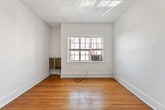 This efficiently designed studio in Birmingham, Alabama offers a well-organized layout across a single floor, comprising a bedroom, kitchen, bathroom, and hallway. The bedroom is the largest space in the unit, featuring warm hardwood flooring, crisp white walls, and a ceiling fan with a light fixture that provides both airflow and illumination. A window along the exterior wall invites natural light and includes a wall-mounted heating and cooling unit beneath it. The room also offers a closet for storage and direct access to the hallway leading to the rest of the unit. The kitchen is a compact galley-style space equipped with white cabinetry, open shelving, a gas range, a single-basin sink with a warm wood-toned countertop, and a full-size refrigerator. Light gray vinyl plank flooring gives the kitchen a clean, practical finish. The bathroom is a standout feature, showcasing white ceramic tile throughout with a bold cobalt blue tile accent band that adds a striking visual element. A deep soaking tub with a shower attachment is framed by a large frosted window that fills the room with soft, diffused natural light. Overall, the space presents a functional and straightforward living environment with classic character details.