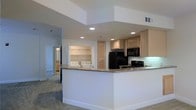 This single-floor residence offers a thoughtfully designed open-concept layout that seamlessly connects the kitchen and living areas. The kitchen is a functional centerpiece, featuring light wood cabinetry, granite countertops, and a suite of black appliances including a refrigerator, gas range, and built-in microwave. Recessed lighting illuminates the space from above, lending a clean, modern feel throughout. A peninsula counter extends outward, providing additional workspace and a natural division between the kitchen and the adjoining living area.

One of the standout practical features of the home is the in-unit laundry alcove, conveniently situated just off the main living space. This dedicated nook houses a washer and dryer side by side and is outfitted with shelving above, maximizing utility within a compact footprint.

The living area is finished with neutral-toned carpet, contributing to a warm and comfortable atmosphere. White walls and crisp baseboards maintain a clean aesthetic throughout, while the open sightlines between rooms create a spacious, cohesive feel. A hallway leading toward additional rooms suggests a well-organized floor plan designed for everyday comfort and ease of living.