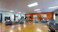 This single-floor community space is thoughtfully designed to foster social interaction and comfortable gathering. The room makes an immediate impression with its bold terracotta-orange accent wall, which contrasts beautifully against the surrounding sage-green and neutral tones, creating a warm and energizing atmosphere throughout.

The space features warm-toned hardwood flooring that extends across the entire area, adding a sense of continuity and natural warmth underfoot. Above, a suspended ceiling is fitted with multiple recessed panel lighting fixtures, ensuring the space is evenly and brightly illuminated throughout. Natural light also filters in through several windows dressed with patterned curtains and white blinds, further enhancing the open and airy feel.

The layout is divided into distinct yet fluid zones. A lounge area is furnished with a contemporary sofa, accent chairs in soft blue upholstery, and a low coffee table arranged atop a layered area rug — ideal for informal conversation and relaxation. Additional seating clusters offer more intimate arrangements for smaller groups. A dining or activity table with chairs sits in the foreground, providing space for meetings or communal activities.

Lush tropical potted plants flank a set of elegant French doors, adding life and texture to the space, while framed artwork adorns the walls, contributing a polished, welcoming character to the overall environment.