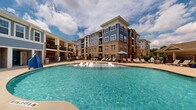 Located at 5000 Green Landing Dr in Cary, NC, this outdoor pool amenity area offers a resort-inspired retreat set within a well-appointed residential community. The centerpiece is a generously sized freeform swimming pool with crystal-clear blue water, gently curved edges, and a gradual depth range clearly marked along the pool's edge. Surrounding the pool is a spacious concrete sun deck lined with an abundance of lounge chairs and chaise lounges, providing ample space for sunbathing and relaxation. A covered pergola-style structure anchors one end of the deck, offering shaded seating and a gathering spot for residents. Additional umbrella-shaded tables are positioned throughout the deck area for outdoor dining and leisure. The pool area is enclosed by a black iron fence with gated access points, enhancing both security and aesthetic appeal. Lush greenery, potted plants, and manicured shrubs are strategically placed to add natural color and privacy. The backdrop of the multi-story residential building, featuring a distinctive combination of blue siding and warm brick, creates an attractive and cohesive architectural setting. Outdoor shower and restroom facilities are accessible directly from the pool deck, adding convenience to this well-designed communal outdoor space.