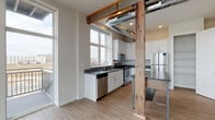 This single-floor loft-style residence masterfully combines industrial design elements with modern, clean finishes throughout. The open-concept kitchen serves as a striking centerpiece, showcasing white shaker cabinetry, dark countertops, a stainless steel sink, and a full suite of stainless steel appliances including a refrigerator, range, microwave, and dishwasher. Rustic reclaimed wood posts and beams contrast beautifully against the exposed metallic HVAC ductwork overhead, lending the space a distinctive character. Light wood-tone flooring flows through the main living and kitchen areas, while large windows fill the space with natural light and offer open outdoor views. A sliding glass door leads to a private balcony with scenic exterior views, accessible from the living area. The bedrooms are carpeted for comfort, each featuring natural light from well-positioned windows. The primary bedroom includes a stylish barn-door entry to a walk-in closet and direct access to an en-suite bathroom. A second full bathroom features a tub-shower combination with tiled surround and wood-tone flooring. An additional half bath with a clean white vanity and mirror rounds out the layout. An in-unit laundry area adds further convenience to this well-appointed space.