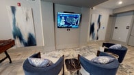 This resident lounge is a thoughtfully designed communal space that blends comfort, entertainment, and sophisticated style. The main seating area anchors the lounge with plush blue accent chairs arranged around a striking black marble coffee table, all positioned on a patterned area rug atop light-toned herringbone wood-look flooring. A wall-mounted flat-screen television serves as the focal point of this gathering zone, complemented by a second mounted TV visible from additional vantage points throughout the space. Large-scale abstract artwork in deep navy and white tones lines the walls, lending a curated, gallery-like atmosphere throughout. A dedicated game room area features a round poker table with upholstered chairs and a foosball table, bathed in natural light from oversized windows dressed with patterned drapes. A high-top glass and steel table with bar-height seating provides an additional spot for casual conversation or casual dining, backed by a textured wood-panel wall art installation. A conveniently placed refreshment station includes a compact refrigerator and countertop service area. Recessed lighting, neutral gray walls, and clean architectural lines tie the entire space together, creating an inviting and functional environment well-suited for both social gatherings and relaxed downtime.