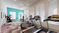 This fitness center is thoughtfully designed to support a comprehensive workout experience within a single, open floor layout. The space is immediately distinguished by its bold teal accent walls, which energize the room and contrast attractively with the neutral white walls and ceilings. Patterned modular carpet tiles in a mix of gray, pink, and teal tones cover the floor throughout, adding both visual interest and functional cushioning underfoot.

Natural light fills the space through multiple windows fitted with white horizontal blinds, creating a bright and welcoming atmosphere. Ceiling fans with recessed lighting provide additional comfort and illumination.

The cardio zone is well-stocked with treadmills, elliptical machines, and a stationary bike, all arranged along the windowed walls to maximize space and airflow. A multi-function cable machine anchors the strength training area, accompanied by a dedicated free weights corner featuring medicine balls, stability balls, resistance bands, and foam rollers neatly stored on a rack. A vertical knee raise and pull-up station adds further bodyweight training options.

Motivational wall art, including a "Keep Calm and Running Every Day" sign, contributes to the encouraging atmosphere. The layout is efficient and uncluttered, offering users a versatile and functional environment for both cardio and strength training workouts.
