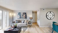 This thoughtfully designed single-floor residence offers a cohesive, contemporary aesthetic highlighted by light hardwood-style flooring, neutral gray walls, and textured ceilings that run throughout the space. The welcoming entryway features a decorative sunburst mirror and a wooden console cabinet, setting the tone for the stylish interiors beyond.

The open-concept living area is bright and inviting, anchored by a large sofa, a dark wood coffee table, and bold wall art. Sliding glass doors allow natural light to pour in while providing access to an outdoor area, creating a seamless indoor-outdoor connection.

The kitchen is efficiently designed with gray cabinetry, a white peninsula countertop, stainless steel appliances including a range, microwave, and refrigerator, along with a tile backsplash. A compact dining area sits adjacent, furnished with a round table and teal accent chairs.

The bedroom is a serene retreat featuring light wood flooring, a tufted upholstered headboard, and a trio of decorative round mirrors above the bed. A vibrant abstract canvas adds a colorful focal point to the room.

The bathroom presents a clean, minimalist design with a floating white vanity countertop, light flooring, and simple chrome fixtures. A built-in closet with bifold doors provides practical storage. The space as a whole balances modern style with functional, comfortable living.