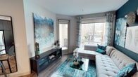 This single-floor residence showcases a cohesive teal and neutral color palette throughout its well-appointed interiors. The living room serves as a welcoming focal point, featuring wood-look flooring, a large statement artwork in oceanic blues, patterned curtains, and a tufted sectional sofa. Corner windows flood the space with natural light and provide views of the outdoor courtyard below.

The kitchen is outfitted with rich dark cabinetry, white countertops, a white tile backsplash, and a full suite of stainless steel appliances including a gas range, microwave, dishwasher, and refrigerator. Recessed lighting and wood-look flooring carry through this space, and a portable island adds additional workspace.

The bedroom features a bold teal accent wall, ceiling fan, patterned window treatments, and a spacious walk-in closet with built-in shelving. The bathroom offers a clean, modern aesthetic with a tub-shower combination, built-in shelving niche, white tile surround, and a dark-toned vanity with white countertop.

The building's exterior features a resort-style pool courtyard with lounge seating, palm trees, and a sweeping multi-story facade — offering residents an elevated outdoor amenity experience.