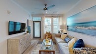 This single-floor residence showcases a bright, coastal-inspired aesthetic throughout, with light wood flooring, crisp white walls, and a soothing blue-and-neutral color palette that creates a relaxed, airy ambiance.

The open-concept kitchen is a standout feature, equipped with white shaker cabinetry, stainless steel appliances, granite countertops, and a large center island with a built-in sink and seating for multiple guests. Pendant lighting overhead adds a modern touch, while a sliding barn-style door provides a distinctive accent.

The kitchen flows seamlessly into the living area, which benefits from abundant natural light through large windows and a glass door leading outside. A ceiling fan overhead and wide-plank flooring enhance the comfortable, laid-back atmosphere.

A dedicated dining nook features a marble-topped table alongside a charming built-in shelving and desk area — ideal as a home workspace or display space.

Both bedrooms are generously sized with plush carpeting, large windows, and ceiling fans. The primary bedroom connects to a well-designed ensuite bathroom featuring dual granite vanities, a glass-enclosed walk-in shower, and a walk-in closet. The second bathroom offers a full tub-shower combination with matching granite vanity and clean tile finishes throughout.