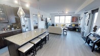This single-floor space showcases a thoughtfully designed open-concept layout that seamlessly connects the kitchen, dining, and living areas into one cohesive and functional environment. The kitchen is a standout feature, anchored by a large granite-topped island with bar seating, offering ample prep space and a natural gathering point. Stainless steel appliances, including a French door refrigerator with an ice and water dispenser, add a polished, contemporary touch, while stylish pendant lighting overhead enhances the modern aesthetic. Light grey luxury vinyl plank flooring runs throughout, tying the space together with a clean, consistent look. Track lighting on the ceiling provides additional illumination, complementing the natural light that pours in through the generous windows in the living area. The living room is spacious and inviting, with room for a full sectional sofa arrangement, a cozy seating corner, and decorative accents including bold wall art and floating shelves. The neutral colour palette of soft whites and greys is accentuated by pops of yellow and teal, creating a lively yet balanced ambiance. Overall, the space strikes an ideal balance between modern design and everyday livability.