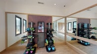 This single-floor fitness center on Bonnar Bridge Pkwy in Burlington, NC, is thoughtfully designed to support a comprehensive workout experience. The main exercise area features light wood-tone flooring and a clean, open layout that provides ample room for movement and training. A prominent accent wall in deep burgundy serves as the focal point of the space, housing an organized rack system stocked with a wide variety of fitness equipment, including kettlebells, resistance tools, medicine balls, weighted bags, and a central interactive fitness display screen. Large floor-to-ceiling mirrors with light wood framing line one wall, visually expanding the space and supporting form-focused training. Multiple black-framed windows are positioned throughout the room, inviting abundant natural light and offering views of the surrounding outdoor greenery. The vaulted ceiling adds a sense of openness and airiness to the workout environment. Adjacent to the main floor, natural wood interior doors lead to a dedicated storage room and additional utility areas, keeping the workout space uncluttered. The overall design strikes a balance between functional fitness infrastructure and a clean, modern aesthetic, making it a well-appointed facility for residents in the Burlington, NC area.