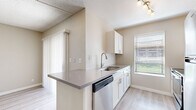 This thoughtfully designed 1-bedroom, 1-bathroom unit offers a functional and modern living experience across a single floor. Light wood-tone vinyl flooring flows consistently throughout, creating a cohesive and clean aesthetic in every room.

The kitchen serves as a central feature, showcasing white shaker-style cabinetry, grey quartz countertops, a peninsula with a breakfast bar overhang, a stainless steel dishwasher, range, and refrigerator, plus a single-basin sink with a gooseneck faucet. A window above the sink fills the space with natural light.

The living room is open and airy, featuring a textured ceiling, built-in wall shelving for display or storage, and neutral warm-beige walls. A hallway connects the main living areas to the private bedroom and bathroom.

The bedroom is generously sized, with a ceiling fan, a large window with exterior views, and access to a dedicated walk-in closet. The closet is well-organized with dual hanging rods and a central shelving unit, offering ample storage capacity.

The bathroom presents a modern, spa-like aesthetic with a floating wood-finish vanity, a framed mirror with vanity lighting, and a combined shower/tub. Wood-look tile flooring and clean white walls complete the refined look.