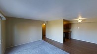 This single-floor space presents an open and functional layout that connects the main living area to the kitchen. The living area features a combination of soft carpet and hardwood-style flooring, offering a practical and comfortable underfoot experience. Neutral-toned walls and a standard white ceiling contribute to a clean, understated aesthetic throughout.

A doorway visible from the living area leads further into the unit, suggesting additional rooms beyond. The kitchen, partially open to the living space, is outfitted with dark wood cabinetry and dark countertops, providing a notable contrast to the lighter walls. An appliance is partially visible beneath the counters, indicating functional kitchen infrastructure. A ceiling vent is present in the main area, pointing to a central HVAC system for climate control.

Natural light appears limited based on the image, with overhead lighting supplementing illumination. The overall ambiance is modest and straightforward, with the open connection between the living and kitchen areas lending the space a sense of flow suited to everyday living.