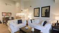 This single-floor space showcases an open-concept design that seamlessly connects the living area to the kitchen. The living room is a standout feature, boasting soaring vaulted ceilings that amplify the sense of openness and airiness throughout the space. Crisp white walls reflect an abundance of natural and ambient light, contributing to the room's clean, bright atmosphere.

The living area is anchored by two generously sized white slipcovered sofas accented with navy and coral-motif throw pillows, evoking a relaxed coastal aesthetic. A dark wood coffee table serves as a central focal point, complemented by a woven area rug beneath. Two dark-framed mirrors are mounted on the wall, adding depth and visual interest to the space.

The kitchen, visible through an open passthrough, features white cabinetry, white appliances, and a raised breakfast bar that provides a casual dining option while maintaining the open feel between rooms. The consistent use of white tones throughout both spaces creates a cohesive, inviting ambiance ideal for comfortable everyday living.
