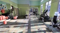 This fitness center offers a bright and welcoming environment designed to support a comprehensive workout experience. The space is finished with vibrant light green walls accented by decorative striping near the ceiling, creating an energizing atmosphere. Multicolored striped carpet tiles cover the floor, adding a lively visual element while providing a comfortable surface underfoot.

Large windows along the perimeter flood the room with natural light, enhancing the open and airy feel of the space. Full-length mirrors line one wall, making the room feel even more expansive while allowing users to monitor their form during exercise.

The facility is outfitted with a solid variety of equipment, including a multi-station cable and strength machine, a row of dumbbells on a dedicated rack, and cardio machines such as an elliptical trainer. Stability balls are also available for functional and core training. A stainless steel trash receptacle is conveniently positioned within the space, reflecting attention to cleanliness and usability.

Access to an adjoining room is visible through a doorway, suggesting additional amenities or utility space. Natural light streams across the floor, highlighting the well-maintained condition of the facility. Overall, this fitness center is a functional, inviting space suited for a wide range of exercise routines.