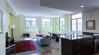 This single-floor space is designed with an open-plan layout that integrates the living and kitchen areas into one cohesive environment. Large windows wrap around the space, flooding the interiors with natural light while framing views of surrounding urban greenery and city buildings. The light-colored walls complement the bright, airy atmosphere throughout.

The living area features hardwood flooring that transitions to carpet, anchored by a bold red area rug that adds a pop of color. A contemporary gray sofa serves as the centerpiece, accompanied by a sleek glass coffee table and a black accent chair, creating a comfortable and stylish seating arrangement. A floor lamp with a cylindrical shade provides additional ambient lighting.

Adjacent to the living area, a dining space accommodates a dark table with modern chairs, seamlessly connecting to the kitchen. The kitchen features a dark, glossy island countertop that extends into the space, offering both a workspace and a visual boundary between the cooking and living zones. The overall design aesthetic is clean and modern, with a neutral palette punctuated by contrasting dark cabinetry and furnishings.