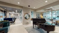 The Piano Lounge is a thoughtfully designed amenity space that seamlessly blends refined elegance with functional versatility. At its heart, a polished black grand piano anchors the entry area, set against light-toned large-format tile flooring and framed by a decorative gold-leaf mirror that adds a touch of glamour to the neutral palette. Glass-walled rooms line the corridor, offering transparency and an open, airy feel throughout.

The lounge area invites relaxation with plush teal velvet seating, layered textures, and artfully curated décor, including sculptural pendant lighting that cascades from the ceiling. Floor-to-ceiling glass doors and windows bathe the space in natural light while providing direct views and access to an outdoor courtyard.

A standout feature is the expansive demonstration kitchen, equipped with a generous white quartz island with bar seating for multiple guests, professional-grade appliances, a stainless steel range hood, and a mosaic tile backsplash that adds warmth and texture. Navy cabinetry grounds the space with a bold contrast against the bright countertops.

Throughout, the design maintains a cohesive modern aesthetic—clean lines, warm neutrals, and carefully chosen accent pieces—creating an inviting environment ideal for social gatherings, culinary events, and everyday community living.