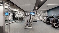 Located at 590 Park St in St. Paul, Minnesota, this well-appointed fitness center offers a comprehensive workout environment across a single open floor. The space is designed with a modern aesthetic, featuring a striking black-and-white city map mural that spans multiple walls, creating a bold and urban visual identity. Complementing the mural, one wall is finished in a deep steel-blue tone, adding depth and contrast to the overall design.

The fitness center is equipped with a full suite of Life Fitness equipment, including treadmills and elliptical trainers with integrated touchscreen displays for cardio training, as well as a selection of strength machines and a dumbbell rack with adjustable benches for free weight exercises. A rowing machine and stationary bike further round out the cardio offerings.

Full-length mirrored walls expand the sense of space and provide functional visibility for form-checking during workouts. Recessed linear LED lighting runs throughout the drop ceiling, ensuring consistent, bright illumination across the entire floor. Durable, multi-toned carpet tiles cover the floor, offering comfort and resilience underfoot.

The entry area includes a water fountain station, sanitation supply dispensers, and access to adjacent restroom facilities, ensuring convenience for all users. The layout is open and spacious, allowing for efficient flow between equipment zones.