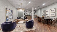 Located at 1514 E Cleveland Ave in East Point, GA, this thoughtfully designed communal lounge space combines comfort and style in a single open-plan floor. The space is anchored by rich hardwood flooring and an eye-catching embossed tin ceiling that lends a distinctive architectural character throughout.

The lounge area is furnished with plush navy velvet swivel chairs arranged around a rustic wood coffee table atop a layered area rug, creating an inviting conversation zone. A large-scale abstract painting and curated framed artwork adorn the white-painted brick and paneled walls, adding artistic depth to the space.

A dedicated dining area features a dark-topped table surrounded by warm wooden chairs, ideal for group gatherings. Nearby, a kitchenette is equipped with dark stone countertops, cabinetry with gold hardware, a sink, and a bold black-and-white geometric tile accent wall that serves as a dramatic focal point.

Recessed lighting and a statement pendant light provide warm, layered illumination throughout. Structural columns are seamlessly integrated into the design. Additional amenities include a foosball table and a large indoor plant. Sage green doors with glass pane details offer access points, completing this polished and functional shared living environment.