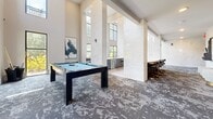 Located in Alpharetta, Georgia, this sophisticated community living room offers a well-appointed, single-floor space designed for both socializing and relaxation. The layout is thoughtfully divided into distinct zones, each with its own character and purpose.

At the heart of the space, soaring double-height ceilings and floor-to-ceiling windows flood the area with natural light, highlighting dramatic marble-clad columns and walls that lend an air of refined elegance. A full-size pool table anchors the game area, complemented by a sleek wet bar with counter seating — perfect for casual gatherings.

A welcoming lounge zone features plush velvet seating in rich navy and blush tones arranged around a marble-top coffee table, with a textured white brick accent wall and a bold dark-tile column supporting a wall-mounted flat-screen TV. Curated abstract artwork adds personality throughout.

A more intimate seating nook near the entrance offers tufted leather benches flanking a pedestal table beside glass doors that open to outdoor greenery, bathed in warm natural light. The space is unified throughout by a striking patterned carpet in grey and charcoal tones, tying together the modern aesthetic. This space delivers a dynamic and stylish environment suited for community events and everyday leisure alike.
