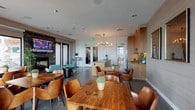 Located at 1011 Washington Blvd in Stamford, CT, the Community Room at Vela on the Park is a well-appointed, single-floor amenity space designed to serve as a versatile hub for socializing and gathering. The room is divided into distinct functional zones, each thoughtfully designed with modern finishes and upscale furnishings.

The lounge area anchors the space with a striking stone-clad fireplace topped by a wall-mounted flat-screen TV. Teal mid-century modern armchairs and a sleek side table create an inviting seating arrangement. French doors open directly to an outdoor terrace, flooding the interior with natural light and providing panoramic elevated views of the Stamford skyline.

The dining area features multiple natural wood tables paired with cognac leather chairs, set against large floor-to-ceiling windows that further showcase the sweeping city views. Warm textured wall treatments and patterned drapery add depth and refinement to this section.

The full kitchen is equipped with stainless steel appliances, including a French door refrigerator, built-in oven, and gas cooktop. Warm wood cabinetry, a stone tile backsplash, and a generous quartz-topped island with a double sink make it fully functional for entertaining. A sculptural multi-globe brass chandelier serves as a focal point overhead.

Large-format tile flooring flows throughout, unifying the space with a clean, contemporary aesthetic.