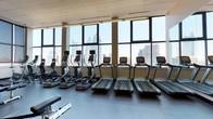 This fitness center offers a comprehensive workout environment across a single open floor plan designed to accommodate a full range of exercise needs. The space is defined by an expansive wall of floor-to-ceiling windows that frames dramatic city skyline views, flooding the interior with natural light and creating an energizing atmosphere for exercise.

The cardio zone lines the window wall and features a row of treadmills and elliptical trainers, each equipped with individual digital display screens. This placement allows users to exercise while enjoying unobstructed views of the surrounding urban landscape. Overhead, recessed ceiling tiles and linear fluorescent lighting provide consistent illumination throughout.

The strength training section is well-appointed with a suite of Cybex cable machines, multi-functional weight stations, and adjustable benches, offering options for both isolation and compound exercises. Full-length mirrors are installed along interior walls, visually expanding the space and allowing users to monitor form. A dedicated functional training area features stability balls, resistance attachments, and flat mats for stretching and core work. A ceiling-mounted television is positioned for easy viewing. Durable rubber flooring throughout ensures safety and sound absorption across all training zones.