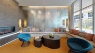 Located at 205–233 E Seaside Way in Long Beach, California, this single-floor space offers a thoughtfully designed interior that blends modern aesthetics with comfortable, functional areas. The main lounge area is anchored by a striking floor-to-ceiling dark tile fireplace surround, complemented by a slatted wood reception bar topped with a white marble-style surface and paired with a round leather barstool. A sculptural multi-arm black pendant light serves as a focal point overhead, while large-format light stone tile flooring extends throughout, adding to the open, airy feel. Curated artwork adorns the crisp white walls, contributing to the gallery-like atmosphere. A separate seating lounge features an expansive built-in sofa set against a dramatic backlit metallic accent wall, accompanied by bold dark wood organic-shaped coffee tables and vibrant blue accent chairs, creating an inviting and design-forward gathering space. Floor-to-ceiling glass walls in another seating nook flood the interior with natural light and offer views of the landscaped outdoor areas beyond. A dedicated vanity room, finished in deep charcoal tones with warm wall sconces and built-in countertop workspace, provides a private and intimate retreat. Together, these areas create a cohesive, upscale environment ideal for community interaction and relaxation.