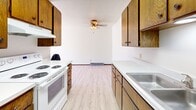 Located at 1502 21st Ave S in Fargo, North Dakota, this one-bedroom, one-bathroom unit offers a practical and efficiently organized single-floor layout. The galley-style kitchen is lined with warm oak cabinetry on both sides, providing generous storage above and below white laminate countertops. White appliances, including an electric range and refrigerator, are integrated neatly into the space, which is finished with light luxury vinyl plank flooring and illuminated by a ceiling-mounted fluorescent fixture. The kitchen opens toward a combined living area that features a ceiling fan with light kit, baseboard heating, and a sliding glass door that leads to a private balcony with outdoor views of surrounding trees and the neighborhood. The spacious bedroom includes a ceiling fan, a closet with bi-fold doors, and a window that brings in natural light. The bathroom is equipped with a tub-shower combination with a handheld showerhead and tiled surround. Throughout the unit, white walls and neutral tones create a clean, bright atmosphere. The subfloor is currently exposed in several areas, indicating the space is in a renovation-ready condition, offering a blank canvas for updated finishes.