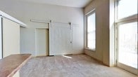 This single-story space is characterized by its clean, neutral aesthetic and practical design elements. The room features soft, wall-to-wall carpeting in a warm gray-beige tone, contributing to a comfortable and understated atmosphere. The walls are finished in a light neutral shade, creating a bright and open feel throughout.

A standout architectural feature is the white sliding barn door mounted on a black metal rail track, which serves as a stylish and space-saving alternative to a traditional hinged door. This detail adds a subtle rustic-modern touch to an otherwise minimalist interior. The doorway it covers provides access to an adjacent area, expanding the functional layout of the space.

Natural light enters the room through a single window fitted with horizontal blinds, allowing for adjustable light control. The window's placement draws light into the corner of the room, brightening the neutral palette. The ceiling height is notably generous, lending a sense of openness and volume to the interior. Overall, the space presents a simple, well-maintained environment with thoughtful design details suited to a variety of uses.