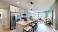 This thoughtfully designed single-floor residence offers a seamless open-concept layout that connects the kitchen, dining, and living areas into one cohesive, light-filled space. The kitchen serves as a central hub, featuring a large island with a white quartz countertop, an undermount sink with a matte black faucet, and bar-height seating. White cabinetry with black hardware pairs with stainless steel appliances, including a French door refrigerator, microwave, and range, while a decorative tile backsplash adds texture. A trio of pendant lights with seeded glass shades hangs above the island, providing both function and style. The living room is anchored by a sage green accent wall adorned with framed artwork and large windows that flood the space with natural light and offer views of greenery outside. A sliding glass door provides access to an outdoor area. Light wood-tone flooring runs throughout the main living spaces, lending warmth and continuity. Both bedrooms are carpeted and generously sized, each featuring large windows and a calm, neutral palette. The primary bathroom offers a dual-sink vanity with quartz countertops, matte black fixtures, a glass-enclosed walk-in shower, and direct access to the bedroom. The secondary bathroom mirrors the same clean, modern aesthetic with a single-sink vanity and bright lighting.