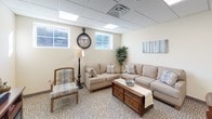 This single-floor facility is thoughtfully designed to provide a comfortable and functional environment for its occupants. The lounge area sets a warm, welcoming tone with plush neutral-toned sofas, a patterned accent chair, and a rustic wooden coffee table arranged around a patterned carpet. Wall art and decorative greenery enhance the relaxed atmosphere, while windows allow natural light to filter in beneath the suspended tile ceiling.

Adjacent to the lounge, the dining area accommodates multiple wooden dining tables and chairs, offering ample seating for group gatherings. Framed artwork adorns the walls, and the space transitions seamlessly into a kitchenette outfitted with light wood cabinetry, a countertop with a sink, a full-size refrigerator, and small appliances including a coffee maker. The kitchen extends into a well-lit hallway with tile flooring that connects the various areas of the facility.

The restrooms are finished to a high standard, featuring earthy stone-look tile covering both the floors and lower walls, grab bars for accessibility, and tasteful decorative accents. The overall design of the space balances practicality with a homey aesthetic, making it well-suited for community use, group activities, or supportive care settings.