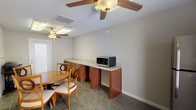 This single-floor space functions as a communal dining or break room, offering a practical and comfortable layout suited for shared use. The room features durable dark tile flooring throughout, complemented by neutral-toned walls with a decorative chair rail detail that adds a touch of architectural character.

At the center of the space sits a round wooden dining table surrounded by matching chairs with upholstered seats, providing a welcoming area for gathering and meals. Along one wall, a built-in counter with warm wood cabinetry and a granite-style surface offers ample workspace, with a stainless steel microwave conveniently placed on top. A stainless steel top-freezer refrigerator occupies a nearby corner, accompanied by a window fitted with horizontal blinds that allows natural light to filter in.

The room is equipped with multiple ceiling fans fitted with light fixtures, ensuring both ventilation and ambient lighting. Fluorescent overhead lighting supplements the ceiling fans, keeping the space well-lit. A standard exterior door provides direct access to the outside. Additionally, a gas grill stored within the room suggests convenient access for outdoor cooking. An illuminated exit sign is also present, consistent with a shared or commercial-use environment. Overall, the space is functional, practical, and well-appointed for communal or organizational use.