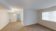 This single-floor residence presents a straightforward, functional layout characterized by clean lines and a neutral color palette throughout. The main living area features wall-to-wall beige carpeting that extends into the connecting hallway, creating a cohesive and comfortable feel underfoot. Bright white walls provide a blank canvas, while a window along one side of the living space allows natural light to filter in, contributing to an airy atmosphere.

A partial wall divides the living area from the adjacent hallway, adding subtle architectural definition without fully closing off the space. The hallway leads toward additional rooms, with a glimpse of a white refrigerator visible near the corridor, indicating the proximity of a kitchen area. A pendant light fixture with a classic design hangs in the hallway, adding a modest decorative touch and supplementing overhead lighting.

The overall ambiance is clean, understated, and move-in ready, with consistent flooring and wall finishes tying the visible areas together. The layout promotes easy navigation between the living space and the rest of the unit, making it a practical and livable environment.