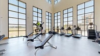 This expansive two-floor fitness center offers a comprehensive workout environment designed to accommodate a wide range of exercise needs. The main floor is the architectural centerpiece of the facility, distinguished by dramatic floor-to-ceiling grid-pane windows that flood the space with abundant natural light and offer open views of the surrounding landscape. Soaring double-height ceilings amplify the sense of openness, while large-format tile flooring lends a clean, modern aesthetic. This primary workout area is equipped with treadmills, elliptical machines, rowing machines, adjustable weight benches, dumbbell racks, and medicine ball storage, providing a full suite of cardio and free-weight options. A dedicated studio room, finished with warm hardwood flooring and an elegant coffered wood ceiling, houses a row of Schwinn spin bikes and features full-length mirrors with ballet barre rails, making it ideal for cycling and movement-based classes. A water station and towel storage are conveniently integrated into the main floor. The upper mezzanine level offers additional strength training equipment, including cable machines and a squat rack, set on textured performance flooring with large windows that maintain the facility's bright, airy character throughout. The overall design balances high functionality with refined architectural detail.