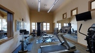This single-story fitness center offers a comprehensive workout environment designed to accommodate a variety of exercise routines. The room features notably high ceilings that contribute to an open, airy atmosphere, while warm beige walls lend a welcoming tone to the space. Natural light filters in through multiple windows along one wall, brightening the interior and providing views of the surrounding greenery outside.

The fitness center is outfitted with a solid selection of equipment, including treadmills for cardio training, a multi-station cable and weight machine for strength exercises, and an adjustable weight bench for free weight workouts. A dedicated dumbbell rack provides a range of free weights, catering to users at varying fitness levels. Protective rubber floor mats are positioned beneath the equipment to ensure safety and floor preservation.

Large wall-mounted mirrors along one side of the room enhance the sense of space while allowing users to monitor their form during workouts. Overhead fluorescent lighting supplements the natural light, ensuring the room remains well-illuminated at all times. The layout is thoughtfully arranged to allow ample space between equipment for comfortable movement throughout the facility.