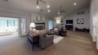 This single-floor clubhouse-style space is designed to serve as a versatile community hub, offering a range of functional and social areas within a cohesive interior. The entry foyer sets an inviting tone with elegant arched doorways, tall ceilings, wall sconces, and patterned carpeting that flows throughout the space. Greenery and tasteful décor reinforce the welcoming atmosphere.

The expansive great room serves as the centerpiece, featuring vaulted ceilings with ceiling fans, recessed lighting, a classic fireplace with a decorative mirror above the mantle, and generous seating arranged around a glass coffee table. A dedicated media area with a wall-mounted flat-screen television offers additional functionality.

Adjacent areas include a lounge with cream leather sectional seating and bold terracotta accent walls, and a dining nook with a round table and black chairs. Large windows throughout provide abundant natural light and direct views of an outdoor pool area. A computer workstation zone with bar-height seating and monitors caters to practical needs. Exposed brick columns and decorative wall art add architectural character throughout, creating a polished and comfortable communal environment.
