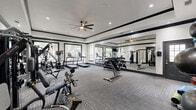 Located at 1000 Farrah Ln in Stafford, TX, this fitness center offers a comprehensive exercise environment designed to support a wide range of workout routines. The single-floor space is thoughtfully laid out to maximize functionality and flow between equipment zones.

The room features a neutral color palette of white and grey, accented by bold dark crown molding that frames the tray ceiling. Recessed lighting throughout ensures a bright, well-lit environment, while a ceiling fan promotes air circulation during intense workouts. Large windows along one wall invite natural light and provide views of surrounding greenery, creating an energizing atmosphere.

A full-length wall mirror visually expands the space and aids in proper form during exercise. The cardio zone includes multiple treadmills and elliptical machines, while the strength training area is equipped with a multi-station cable machine, adjustable weight bench, and a full rack of dumbbells. A dedicated functional fitness area houses stability balls, medicine balls, resistance bands, and a balance trainer.

The flooring consists of durable, striped carpet tiles suited for a gym environment. A wall-mounted television provides entertainment during workouts. An eye-catching motivational mural featuring fitness-themed typography adds personality and energy to the space, reinforcing an atmosphere focused on health and wellness.