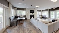 This single-floor space showcases a thoughtfully designed open-concept layout that seamlessly connects the kitchen and main living area. The kitchen is anchored by a curved island with a dark countertop, complemented by white cabinetry, stainless steel appliances, and brass pendant lights suspended from the ceiling. Wood-look flooring flows consistently throughout, reinforcing the cohesive aesthetic.

The expansive lounge area is well-appointed with multiple seating arrangements, including a large sectional sofa and modular banquette-style seating, all centered around a marble-topped coffee table with gold accents. A wall-mounted television sits above a sleek fireplace, creating a natural focal point. Neutral gray walls contrast with bright white trim and drapery, lending the space a polished, contemporary feel.

Multiple windows fitted with blinds and sheer curtains allow generous natural light to fill the room, while French doors open to an exterior deck, enhancing the connection to the outdoors. A separate seating nook near the entry provides additional flexible space. Recessed and skylight-style ceiling fixtures supplement natural lighting throughout. The overall ambiance is modern and inviting, with a refined yet comfortable character suited for communal or multi-purpose use.
