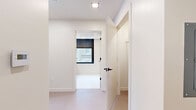 Located on Baltimore Ave in the heart of Kansas City, MO, this efficiently designed unit offers a clean, modern aesthetic throughout its single-floor layout. The open kitchen is outfitted with flat-front white cabinetry extending to the ceiling, a marble-style tile backsplash, stainless steel appliances including a refrigerator, dishwasher, and built-in microwave, and a undermount sink — all contributing to a polished, contemporary feel. Large black-framed windows flood the kitchen and living area with natural light while framing striking views of the Kansas City downtown skyline. The light-toned flooring runs seamlessly throughout, reinforcing the space's cohesive, minimalist design. The hallway connects the rooms efficiently, featuring recessed lighting and crisp white walls that maintain the clean visual language of the space. The bathroom is a standout, showcasing large-format marble-look floor tiles, a warm wood-toned vanity with an integrated sink, and a generously sized black-framed window with roller shade that brings in additional natural light and urban views. Throughout the unit, black hardware accents provide a modern contrast against the predominantly white palette, creating a sophisticated and well-curated interior ideal for urban living.