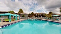 Located at 7000 University Blvd in Winter Park, Florida, this outdoor amenity area offers a resort-style pool experience in a lush, tropical setting. The centerpiece is a generously sized swimming pool with vivid turquoise water, clearly marked depth indicators, and a lane divider, making it suitable for both leisure swimming and lap use. The pool is enclosed by a white metal safety fence, ensuring a secure and well-defined perimeter.

Surrounding the pool, a spacious paver deck finished in warm earth tones provides ample room for lounging, with numerous reclining chairs and chaise lounges positioned along the water's edge. Teal-accented cabanas with draped curtains and coordinating umbrella shade structures offer shaded retreats throughout the deck, creating comfortable spaces to relax out of the Florida sun.

Mature palm trees and lush tropical landscaping frame the area, enhancing the open-air, resort-like ambiance. The backdrop features a two-story residential building with covered balconies and arched architectural detailing, adding a sense of character to the setting. Under the expansive Florida sky, this pool area delivers a vibrant outdoor lifestyle experience ideal for warm-weather relaxation and recreation.