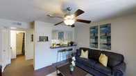 This single-floor residence offers a thoughtfully designed open-concept layout that seamlessly connects the living area to the kitchen. The living room is a comfortable and inviting space, anchored by rich hardwood-style flooring and crisp white walls that create a bright, clean backdrop. A five-blade ceiling fan with an integrated light fixture hangs overhead, providing both airflow and ambient lighting beneath a textured ceiling. The space is accented with contemporary wall art, adding a pop of color and personality to the room.

The kitchen connects directly to the living area via a raised breakfast bar finished with a dark countertop, complemented by matching bar stools — ideal for casual dining or entertaining. A large mirror above the bar reflects light and adds a sense of depth to the space. The kitchen itself features dark cabinetry and modern fixtures, giving it a sleek, polished look.

Throughout the residence, the open flow between rooms creates a sense of spaciousness, making it easy to move between living and dining areas. The overall ambiance is modern and welcoming, with a neutral color palette punctuated by warm wood tones, making it a versatile space suited to a variety of interior styles.