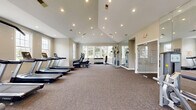 Located at 6460 Chelsea Glen Dr in Columbus, OH, this well-equipped fitness center offers a generous, open floor plan designed to support a comprehensive workout experience. The space is defined by its striking vaulted ceilings with recessed lighting, which create an airy, expansive atmosphere throughout. Large multi-pane windows line the walls, flooding the interior with abundant natural light and offering pleasant views of the surrounding greenery.

The exercise room is outfitted with a robust selection of equipment to accommodate a wide range of fitness routines. Cardio options include multiple treadmills, elliptical machines, and stationary bikes, all arranged to maximize floor space and usability. Strength training is supported by a selection of selectorized weight machines and a dumbbell rack. A wall of floor-to-ceiling mirrors visually expands the space while allowing users to monitor their form during exercise.

Neutral-toned carpet covers the floor, providing a comfortable and practical surface underfoot. A wall-mounted flat-screen television adds an entertainment element to the cardio area. The space is accessed through glass doors and includes clearly marked emergency exits, reflecting thoughtful safety planning. The overall design is clean, modern, and functional, making this fitness center a well-rounded amenity for active individuals.