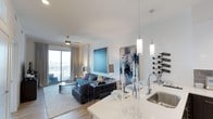 This single-floor residence showcases a thoughtfully designed open-concept layout that connects the kitchen, dining, and living areas into one cohesive, flowing space. The kitchen features a sleek white quartz countertop island with an undermount stainless steel sink, complemented by elegant cylindrical pendant lights that hang overhead, casting warm, focused illumination across the workspace. A wall-mounted wine rack adds a sophisticated, functional accent near the kitchen's edge.

The dining area transitions naturally into the living space, where light wood-toned flooring and neutral-toned walls create a clean, modern backdrop. Bold, large-scale abstract artwork in deep blues and purples serves as striking focal points along the walls, adding artistic character to the space. The living area is anchored by a plush sectional sofa and a contemporary geometric coffee table set atop a textured area rug.

A ceiling fan with integrated lighting sits above the living area, contributing to both comfort and style. Floor-to-ceiling sliding glass doors lead to a private balcony, allowing generous natural light to flood the interior while offering elevated outdoor views. The overall ambiance is polished, contemporary, and welcoming.