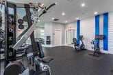 Located at 245 Atlantic Ave in Long Branch, NJ, this well-appointed fitness center offers a bright, thoughtfully designed workout environment across a single open floor. The space is divided into distinct functional zones, each equipped to support a comprehensive exercise regimen.

The cardio area is bathed in natural light from a generous arrangement of large windows and a vaulted ceiling with an arched transom, creating an airy, open atmosphere. Treadmills, an elliptical, and a multi-station cable machine are positioned to take full advantage of the views and light. A wall-mounted flat-screen TV adds entertainment for cardio sessions.

A dedicated free weights section features a full rack of dumbbells, an adjustable bench, and a stretch mat, complemented by a floor-to-ceiling mirror and additional windows that keep the area bright and spacious.

The cardio equipment zone showcases a standout design element — a sculptural three-dimensional textured accent wall adorned with bold vertical blue columns, adding a dynamic and modern aesthetic. Stationary bikes and a rowing machine are thoughtfully arranged in this section, with a wall-mounted TV nearby.

Throughout, durable rubber flooring in a speckled finish ensures safety and performance. Recessed lighting, soft gray walls, and clean white trim tie the entire space together, resulting in a polished and motivating fitness environment.