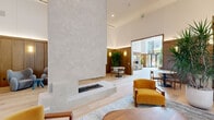 This expansive single-floor space is defined by its sophisticated, biophilic design language, seamlessly blending warm natural materials with contemporary architecture. The centerpiece of the main lounge is a striking, floor-to-ceiling plaster fireplace with a double-sided hearth set on a clean plinth base, anchoring the generous open-plan layout beneath soaring ceilings with cove lighting. Light oak hardwood flooring runs throughout, complementing the warm wood-paneled walls detailed with integrated sconce lighting.

Multiple distinct seating zones furnish the space, featuring an eclectic yet cohesive mix of upholstered sofas, leather lounge chairs, and sculptural accent seating in mustard, terracotta, slate blue, and cream tones. Floor-to-ceiling glazing floods key areas with natural light and frames views of the surrounding streetscape, while sheer drapery softens the light in quieter lounge alcoves.

A dedicated conference or meeting room offers a long wood dining table surrounded by leather chairs, lined with built-in shelving and vertical timber screen partitions. A reception desk area with a green-veined marble counter and vertical wood slat dividers adds refined texture near the entry corridor. Tall, lush tropical plants are thoughtfully placed throughout, reinforcing the vibrant, nature-forward ambiance of the entire space.